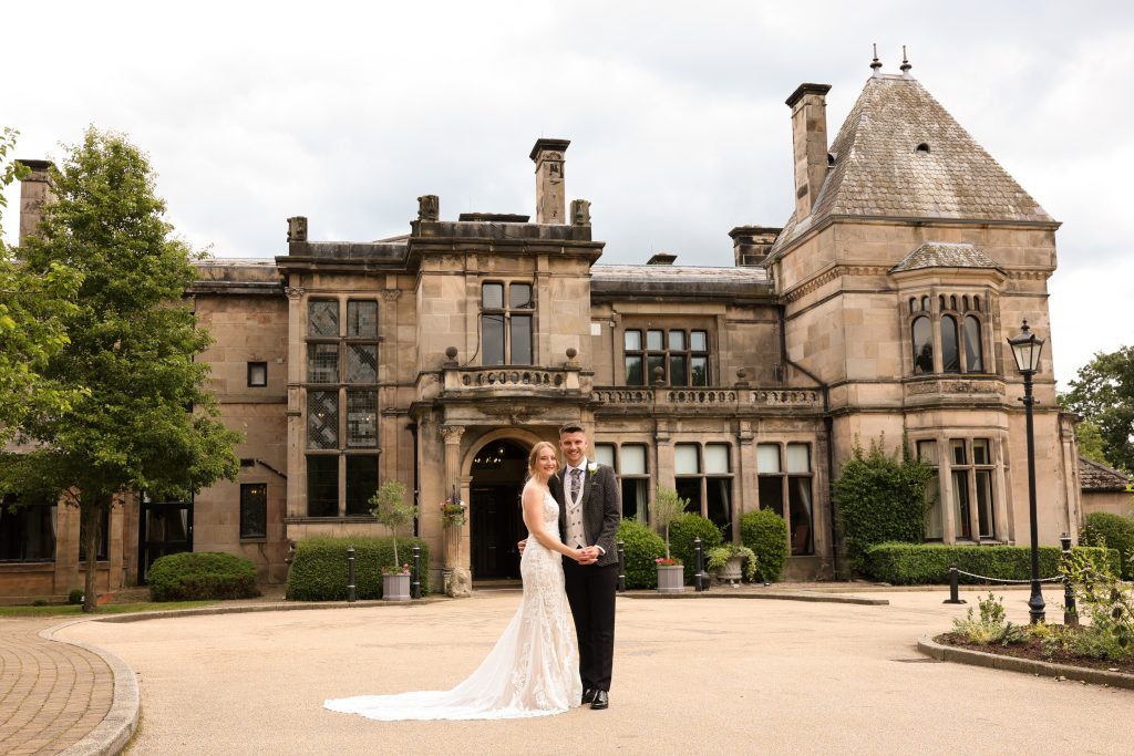 Bride and groom standing posed in front of the stately, grand facade of Rookery Hall, highlighting the classic architecture.