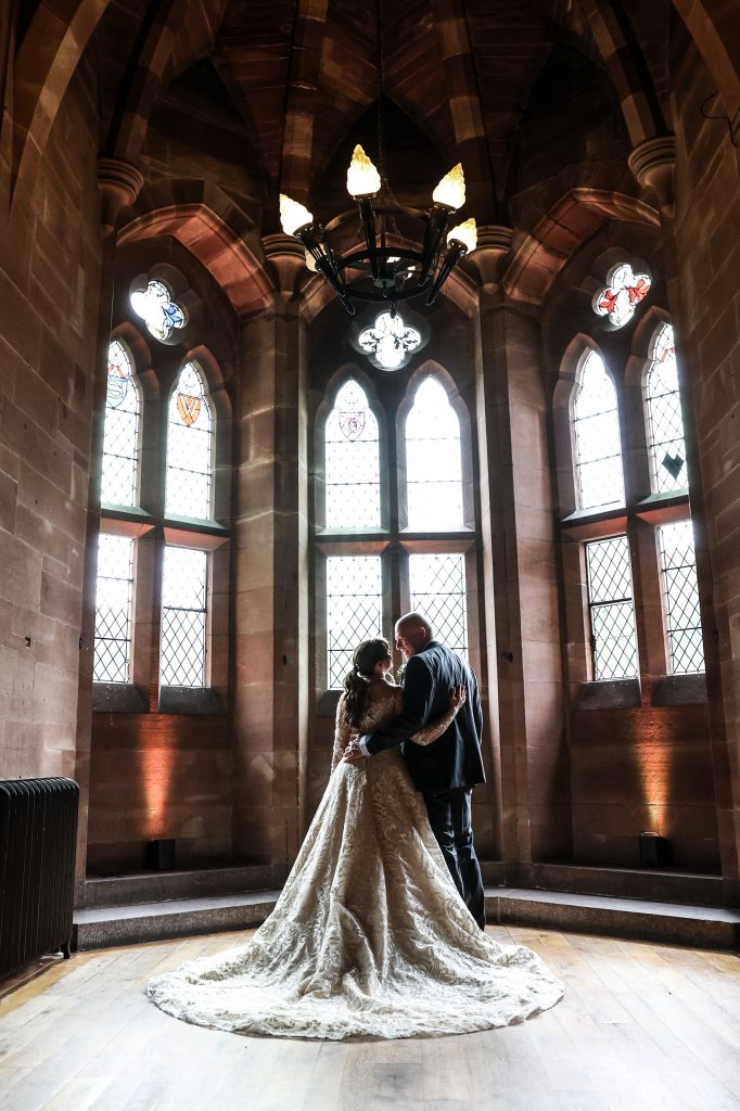 Bride and groom standing intimately framed within the gothic window of the Great Hall at Peckforton Castle.