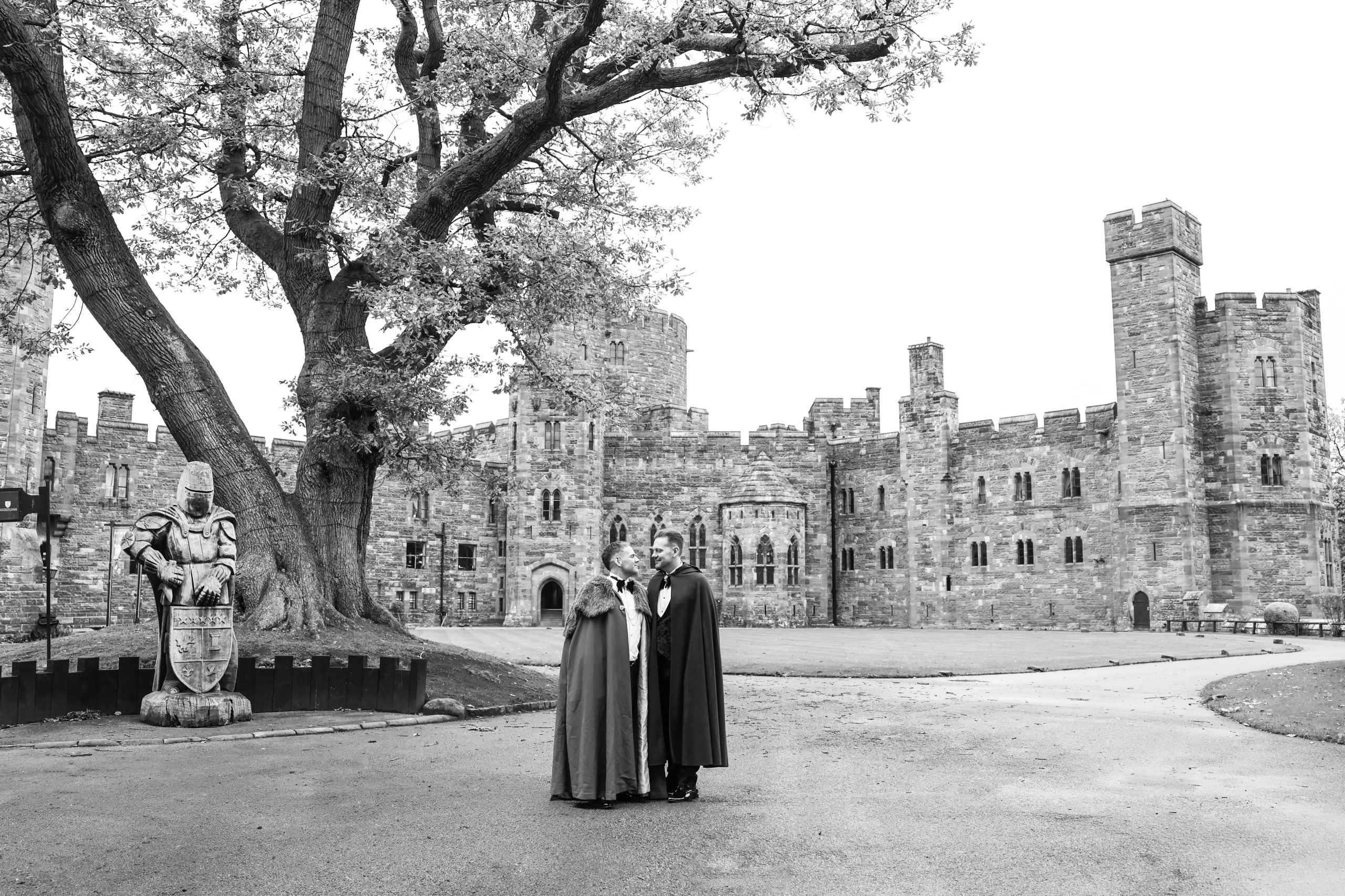 Black and white image of two grooms in sweeping wedding cloaks, embracing in front of Peckforton Castle.