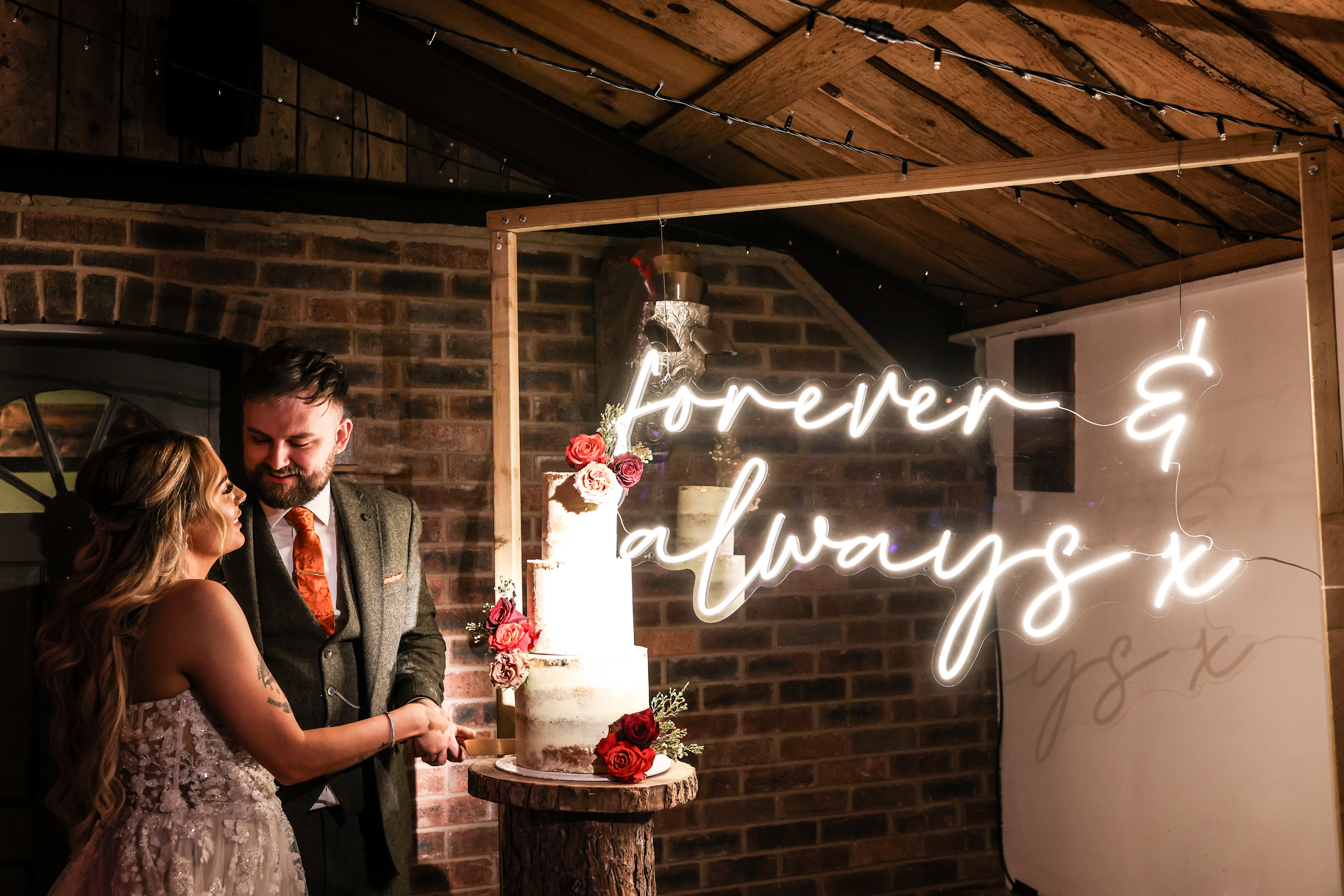Bride and groom cutting their wedding cake in the Lighting Gallery at Owen House Wedding Barn, illuminated by a 'forever & always' neon sign