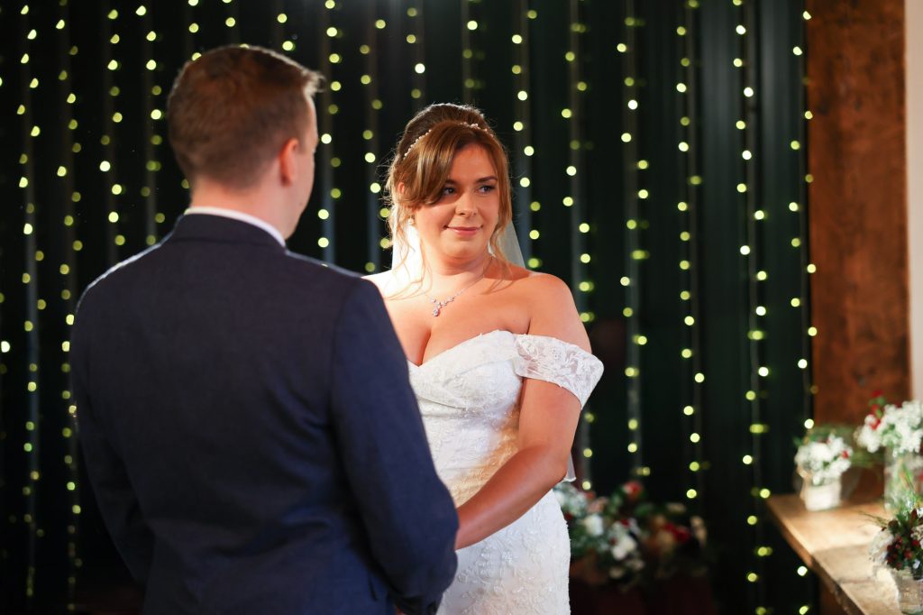 Bride and groom exchanging vows in the Ceremony Barn at Owen House Wedding Barn, framed by soft fairy light backdrop