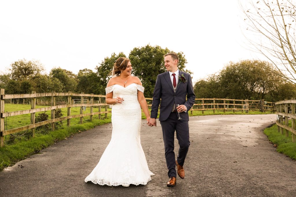 Bride and groom walking hand-in-hand with champagne glasses along a country lane at Owen House Wedding Barn