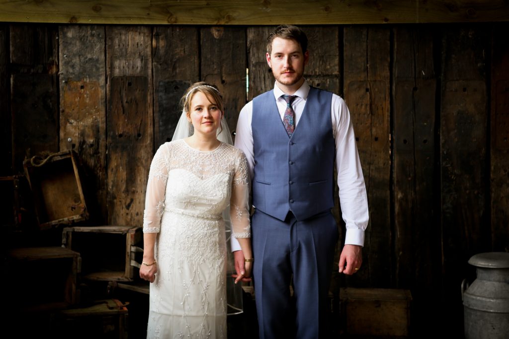 Bride and groom standing hand-in-hand against a rustic wooden barn wall at Owen House Wedding Barn, captured in an editorial portrait style