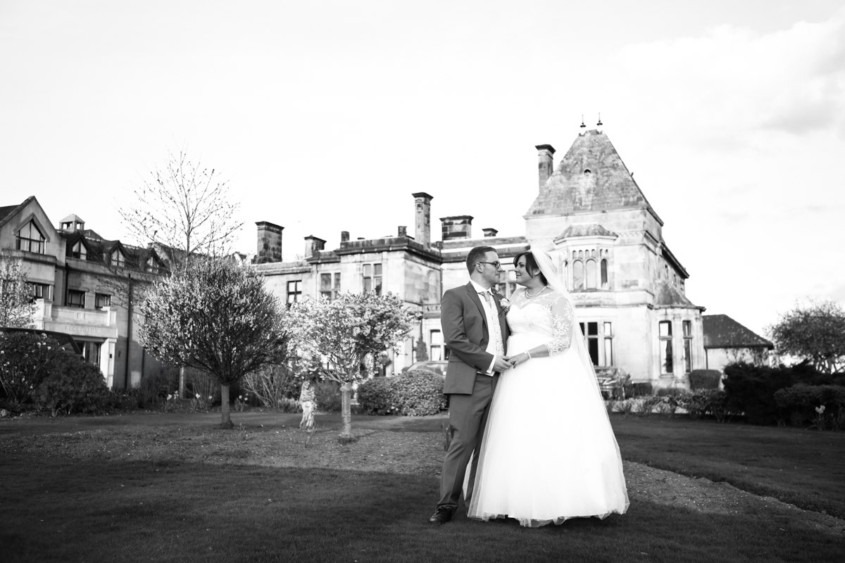 Black-and-white photograph of a bride and groom standing on the lawn in front of Rookery Hall.