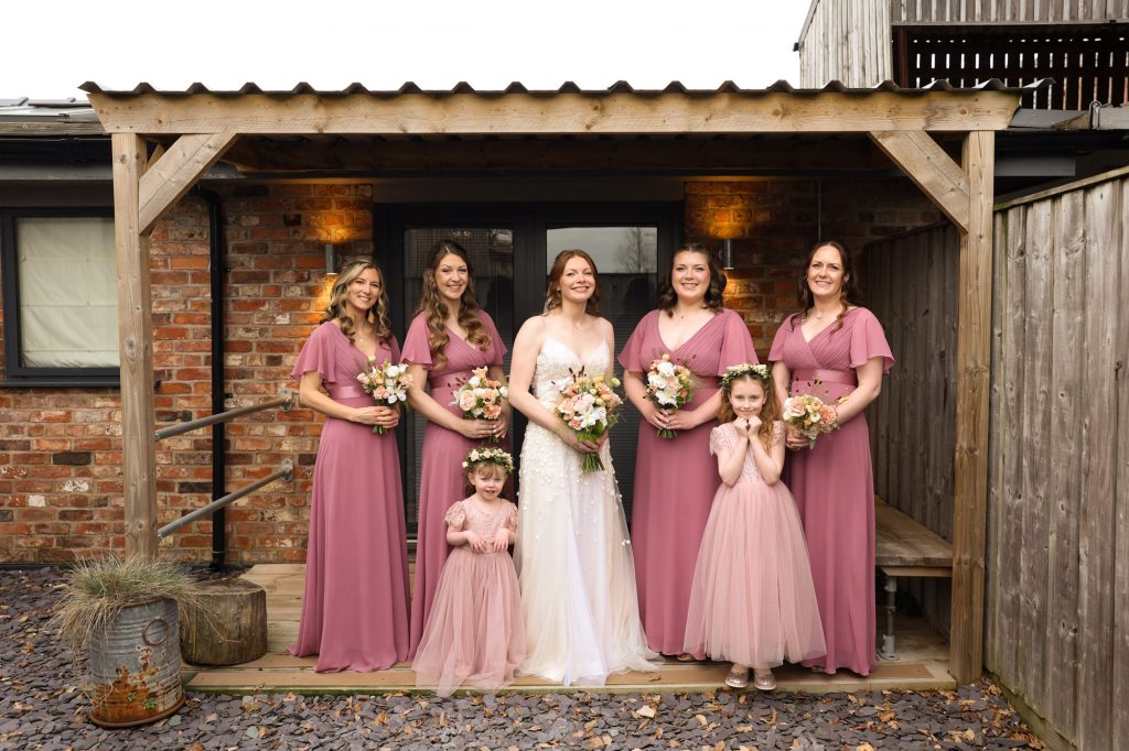 The bride, wearing a white floral-detailed wedding dress, stands with her bridal party outside The Barn Owl House; bridesmaids wear matching dusty rose floor-length gowns, and two flower girls wear complementary pink dresses, all holding bouquets