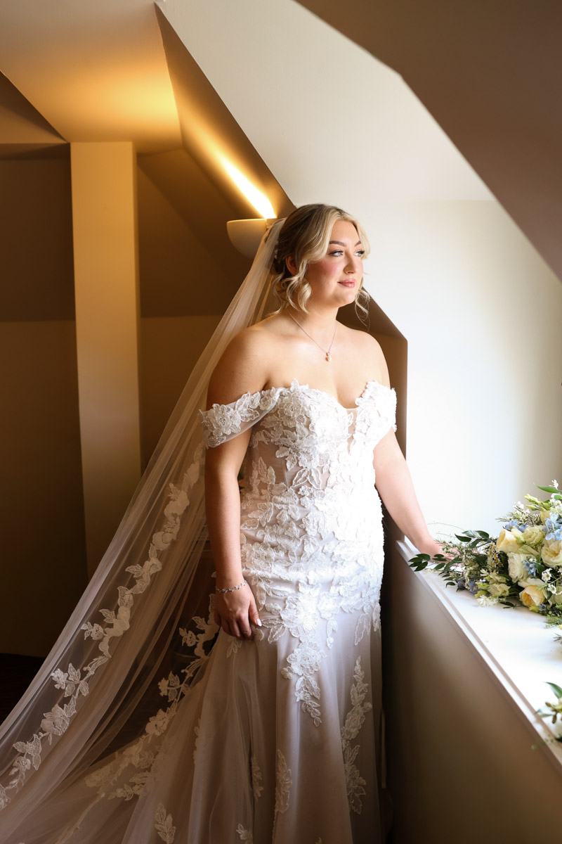 Elegant portrait of a bride in an off-the-shoulder lace gown standing by a window at Rookery Hall.