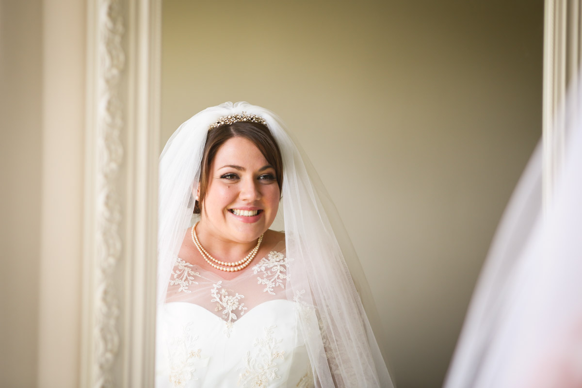 Smiling bride wearing a veil and pearl necklace looking into a mirror during bridal preparations at Rookery Hall.