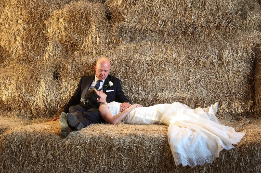 Newlywed couple relaxing together on a stack of hay bales at The Barn Owl House; the bride in a white lace wedding dress rests her head on the groom’s lap, wearing a navy suit