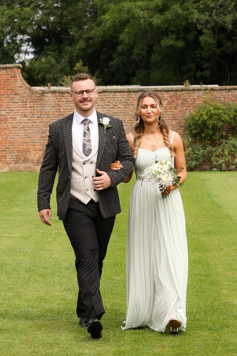 Groomsman and bridesmaid walking arm in arm across the lawn within the ceremony grounds at Rookery Hall.