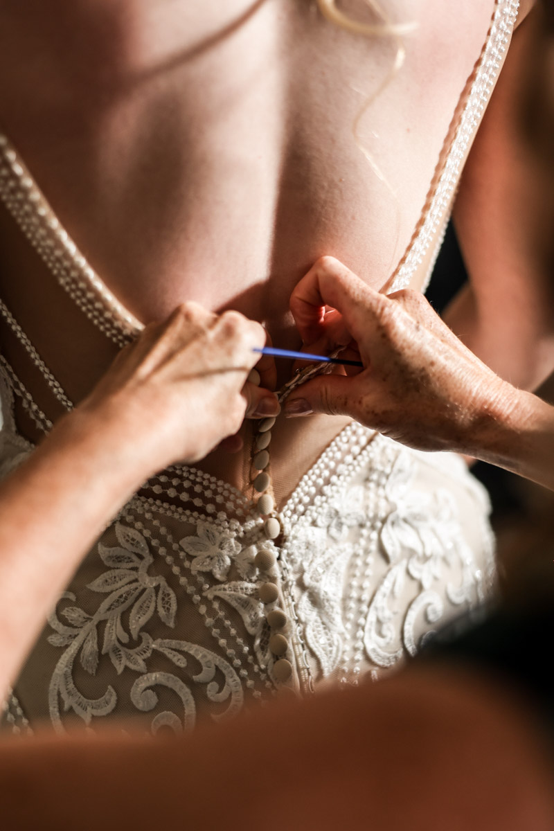 Close-up of hands fastening the buttons on the back of a bride's lace dress during bridal prep at Rookery Hall.