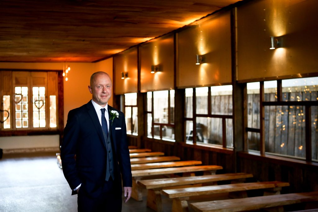 A portrait of a smiling groom in a navy suit, grey waistcoat, and white boutonniere, standing in the aisle of the rustic Barn Owl House wedding barn. Rows of wooden benches line the space, illuminated by warm wall sconces and natural light from large windows overlooking a courtyard