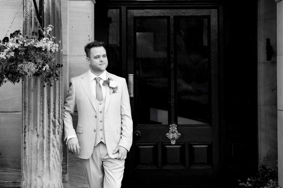 Black-and-white portrait of the groom posing outside a dark wooden doorway at Rookery Hall.