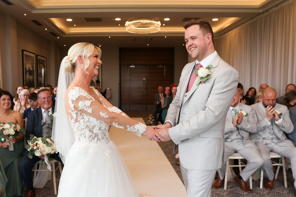 Bride and groom smiling and holding hands during an indoor wedding ceremony at Rookery Hall.
