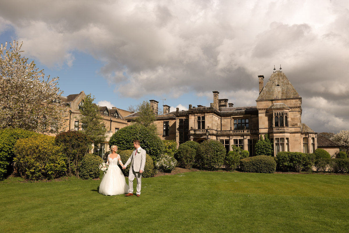 Bride and groom walking hand in hand on the front lawn with the historic facade of Rookery Hall in the background.