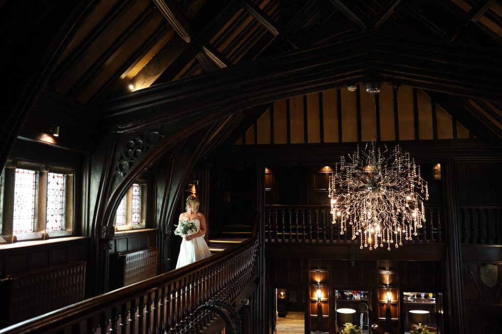 Bride holding a bouquet on the dark wood balcony of Mitton Hall beneath a striking chandelier.
