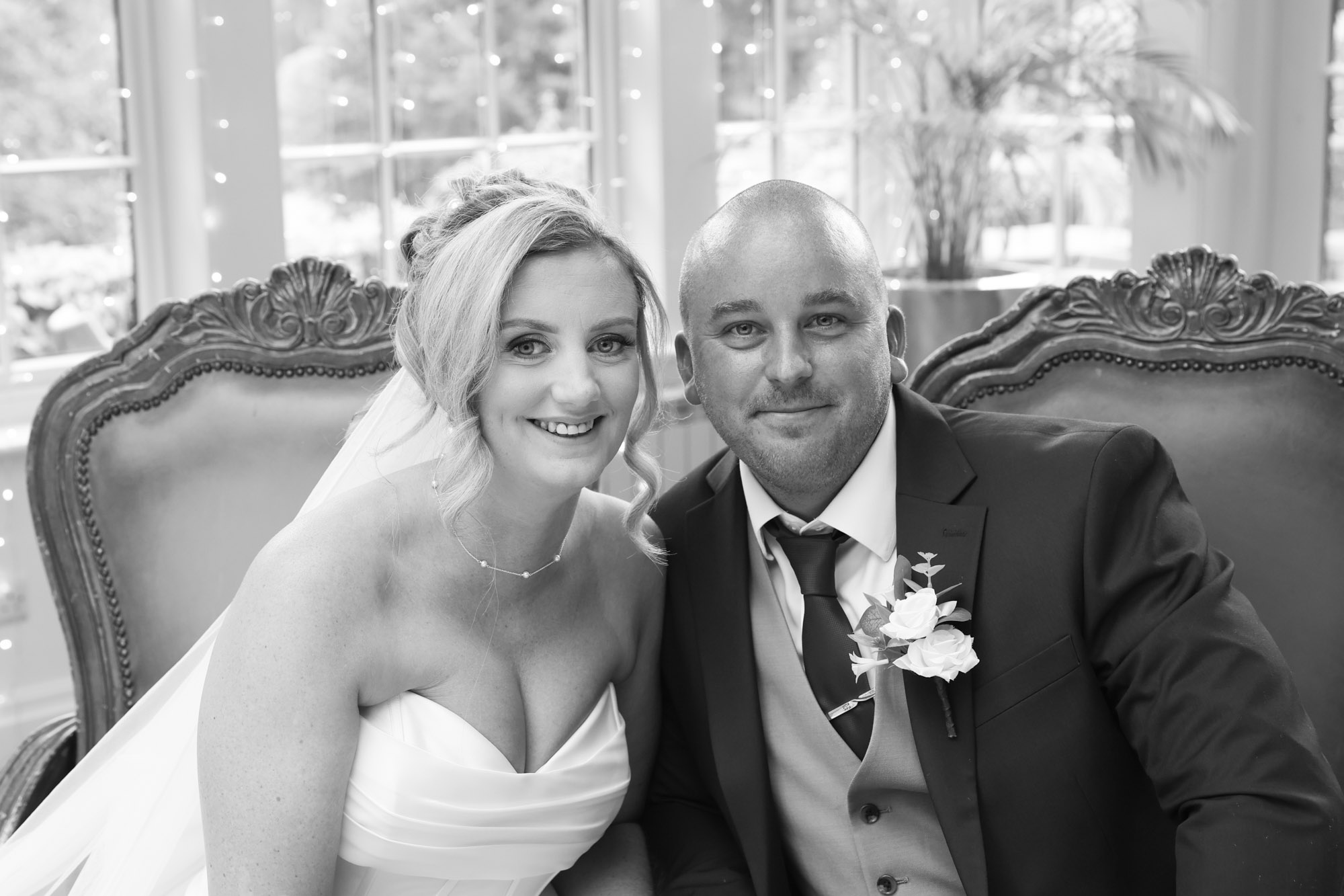 Black and white close-up portrait of the bride and groom smiling together seated on ornate chairs at Mitton Hall.