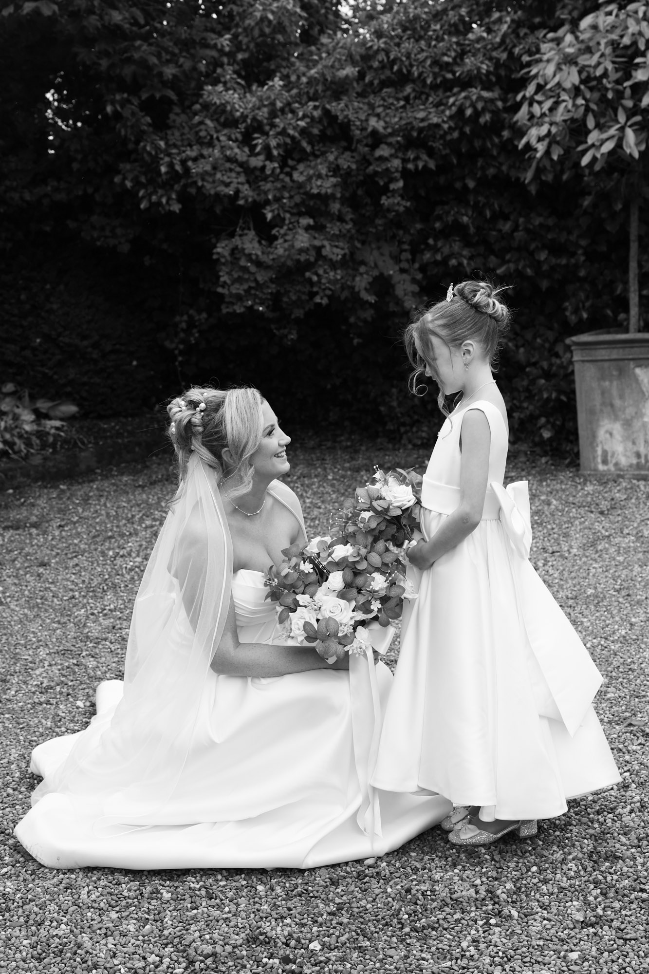 Black and white photo of the bride kneeling down to look at her young daughter, who is holding a bouquet of flowers outdoors at Mitton Hall.