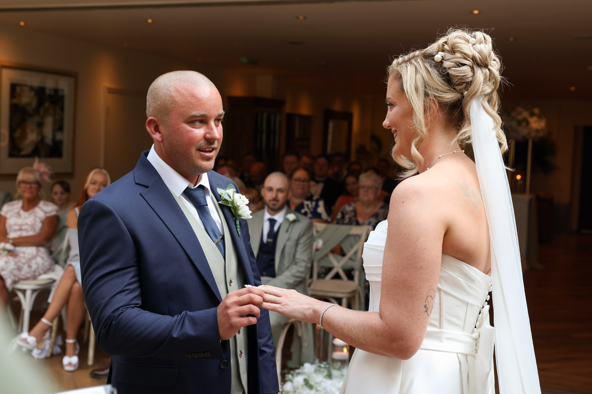 The groom placing a ring on the bride's finger during the ceremony in front of seated guests at Mitton Hall.