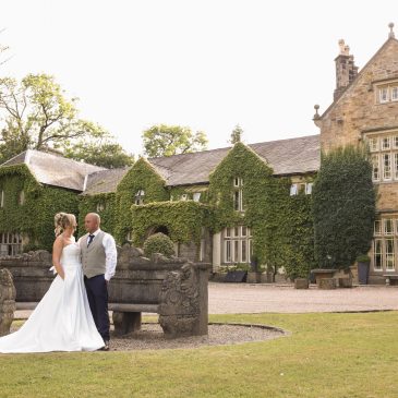 The bride and groom posing together in front of the classic stone bench and ivy-covered exterior of Mitton Hall.