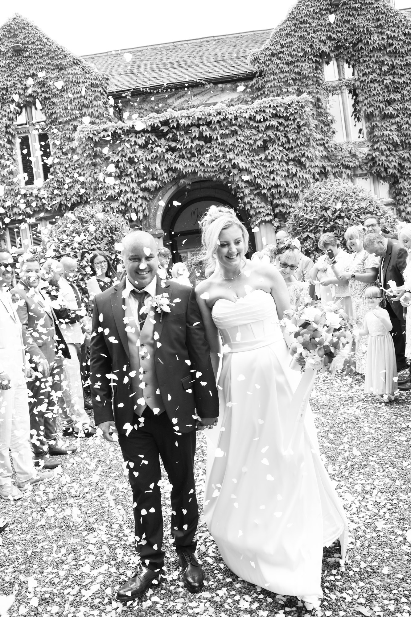 Black and white photo of the bride and groom walking through a flurry of confetti outside the ivy-covered entrance at Mitton Hall.