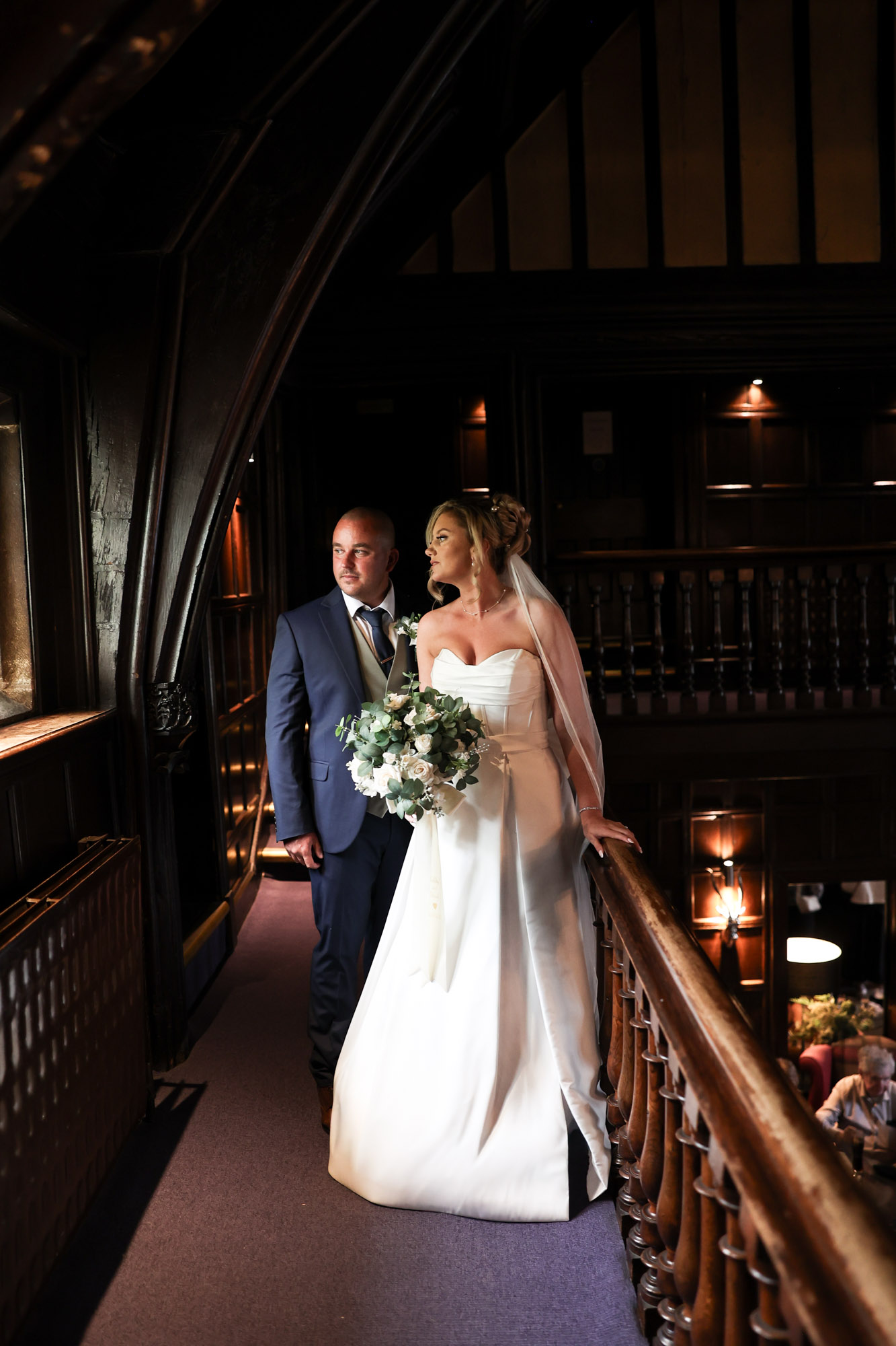 The bride and groom posing together on the dark wood balcony walkway overlooking the interior at Mitton Hall.
