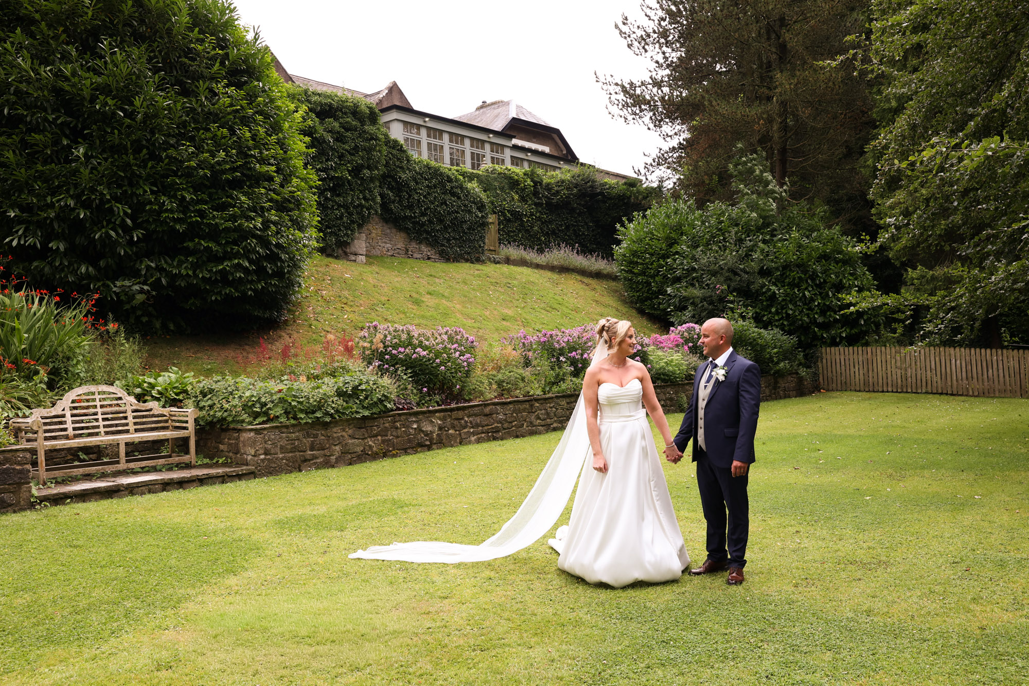 The bride and groom holding hands and looking at each other on the lawn with garden landscaping in the background at Mitton Hall.