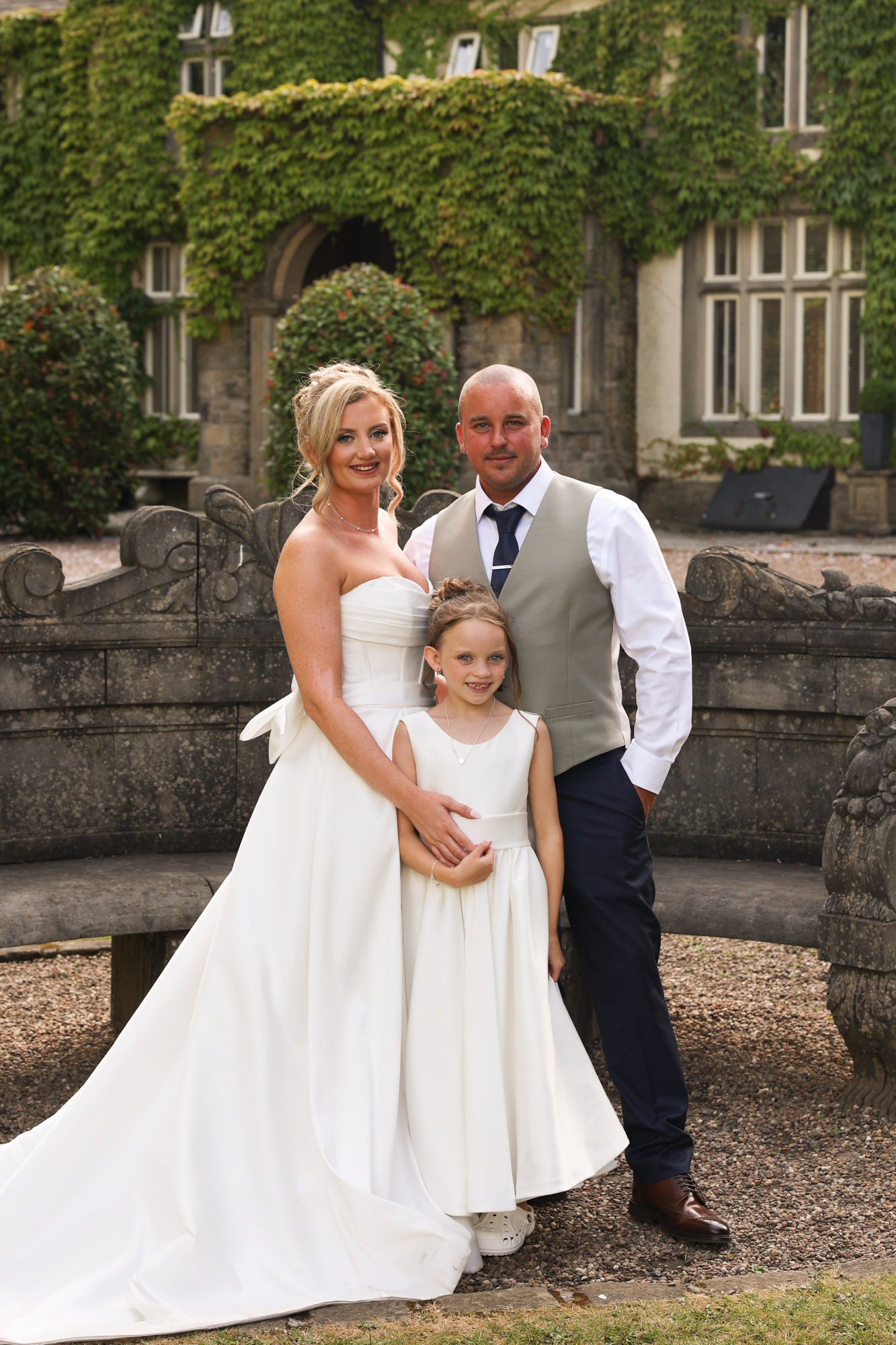 The bride, groom, and young bridesmaid posing together in front of the stone seating and ivy-covered exterior at Mitton Hall.
