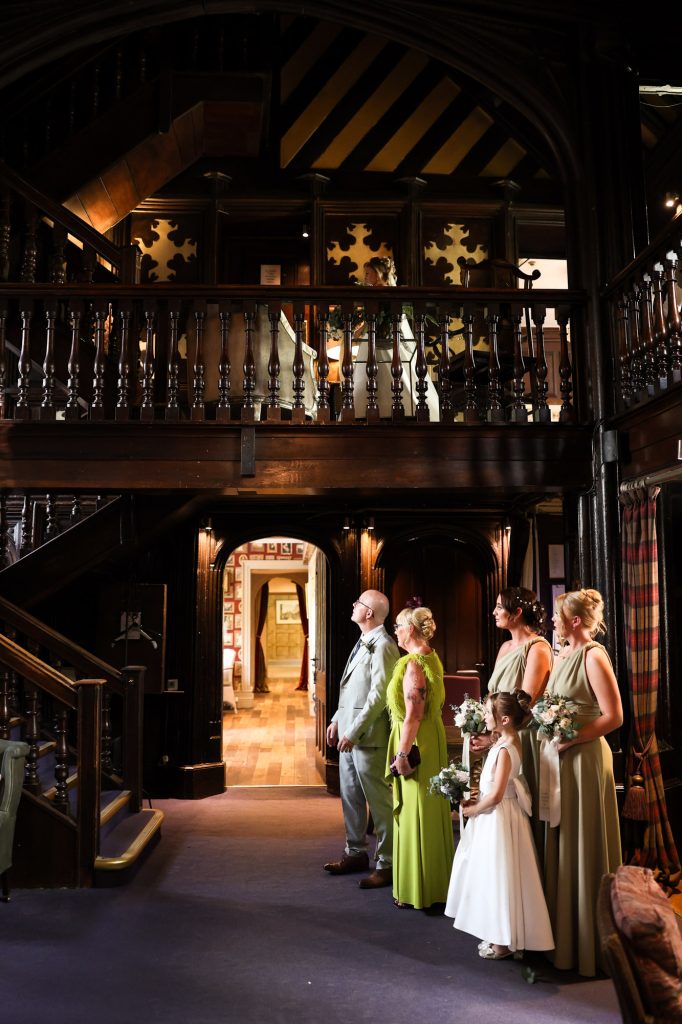 The wedding party looking up from the ground floor as the bride stands on the dark wood balcony above during a first look at Mitton Hall.