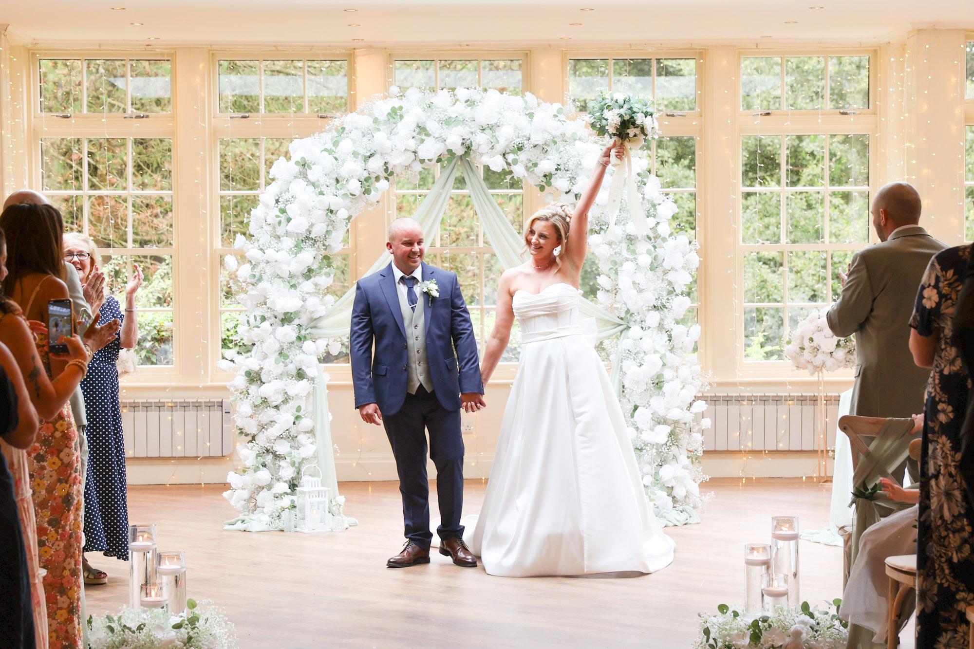 The bride and groom celebrating under a large white floral arch at Mitton Hall.