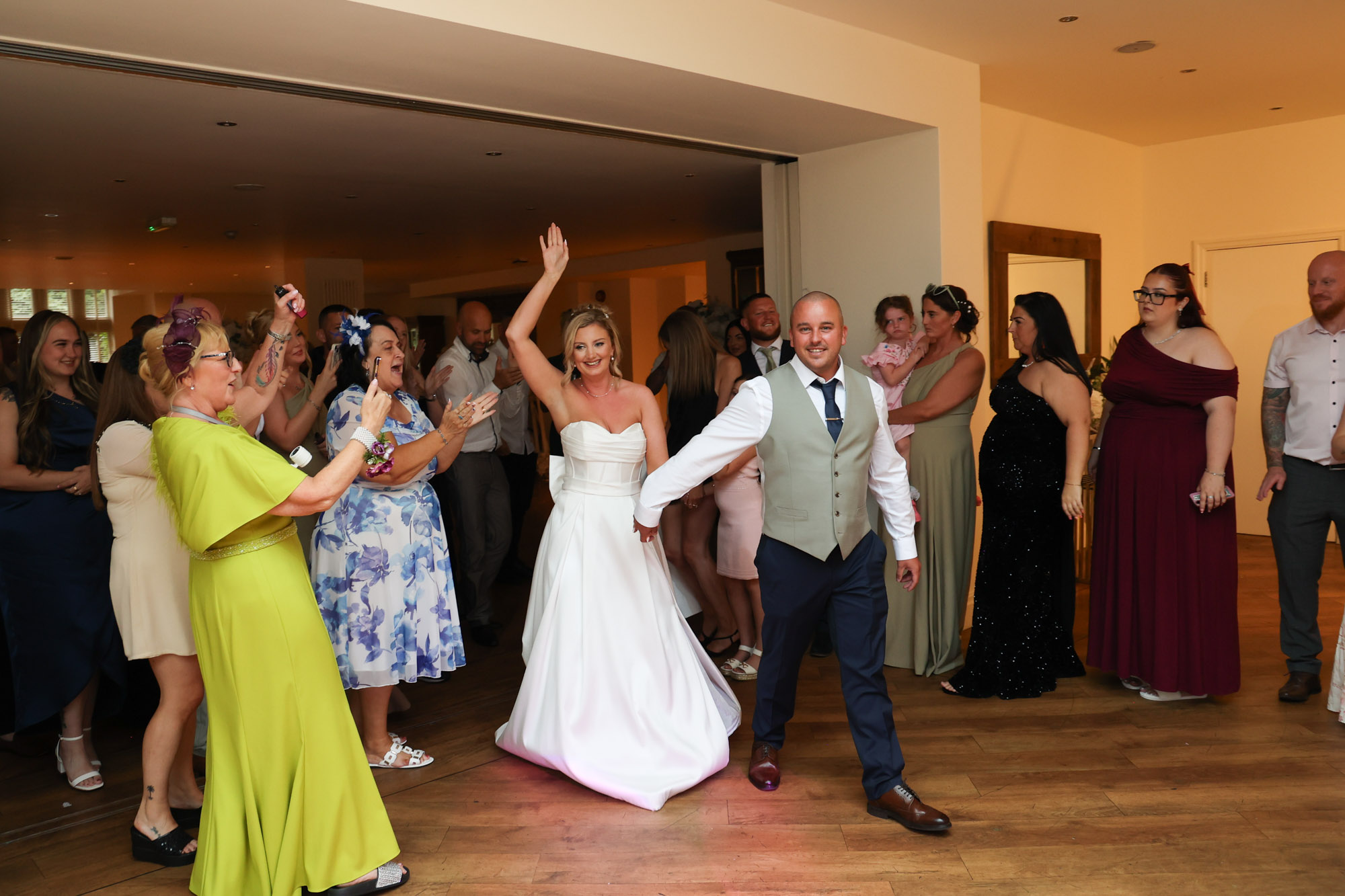 The bride waving with her arm raised as she enters the reception with the groom at Mitton Hall.