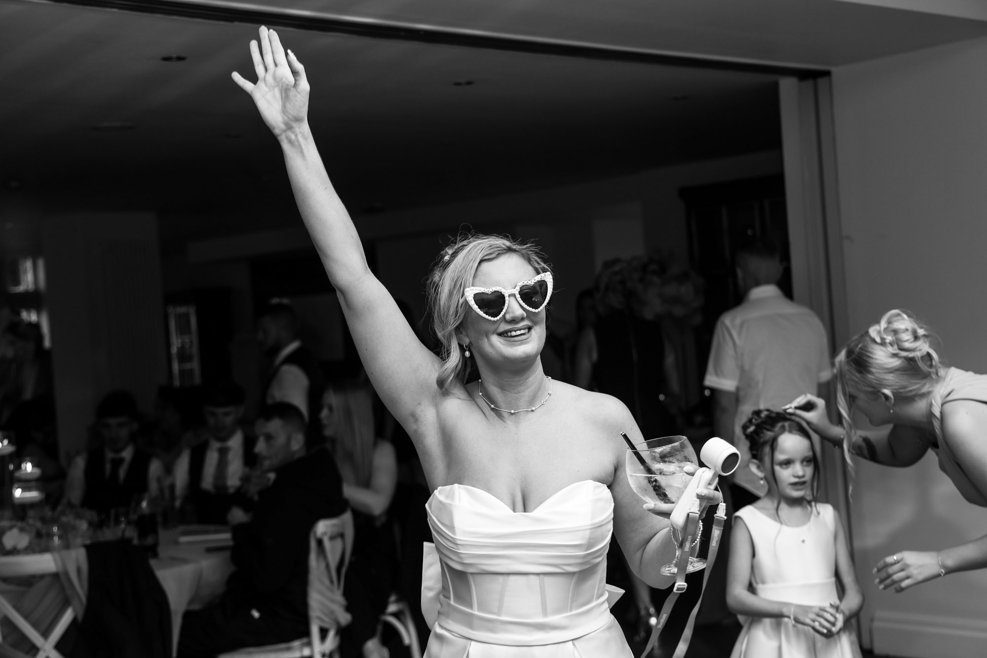 Black and white photo of the bride wearing heart-shaped sunglasses, waving with a drink at the evening party at Mitton Hall.