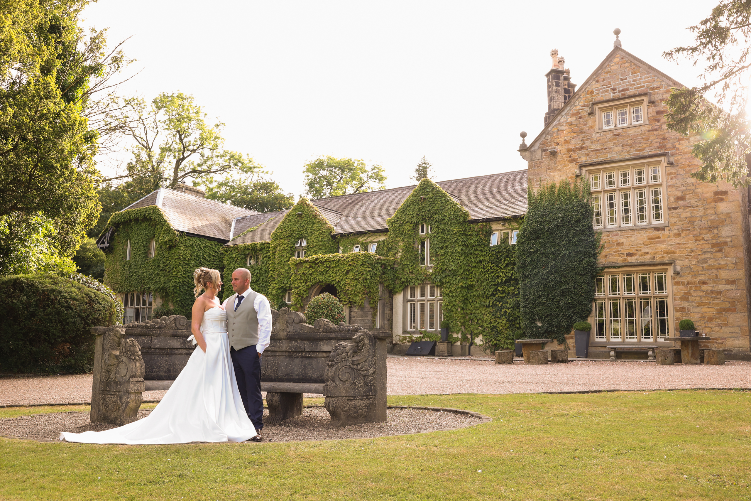The bride and groom posing together in front of the classic stone bench and ivy-covered exterior of Mitton Hall.