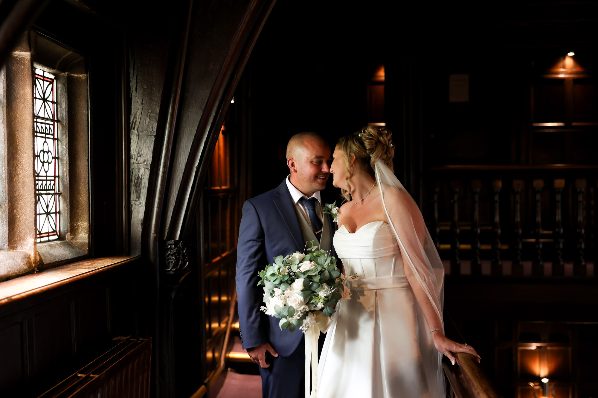 The bride and groom sharing a romantic moment on the dark wood balcony near a stained-glass window at Mitton Hall.