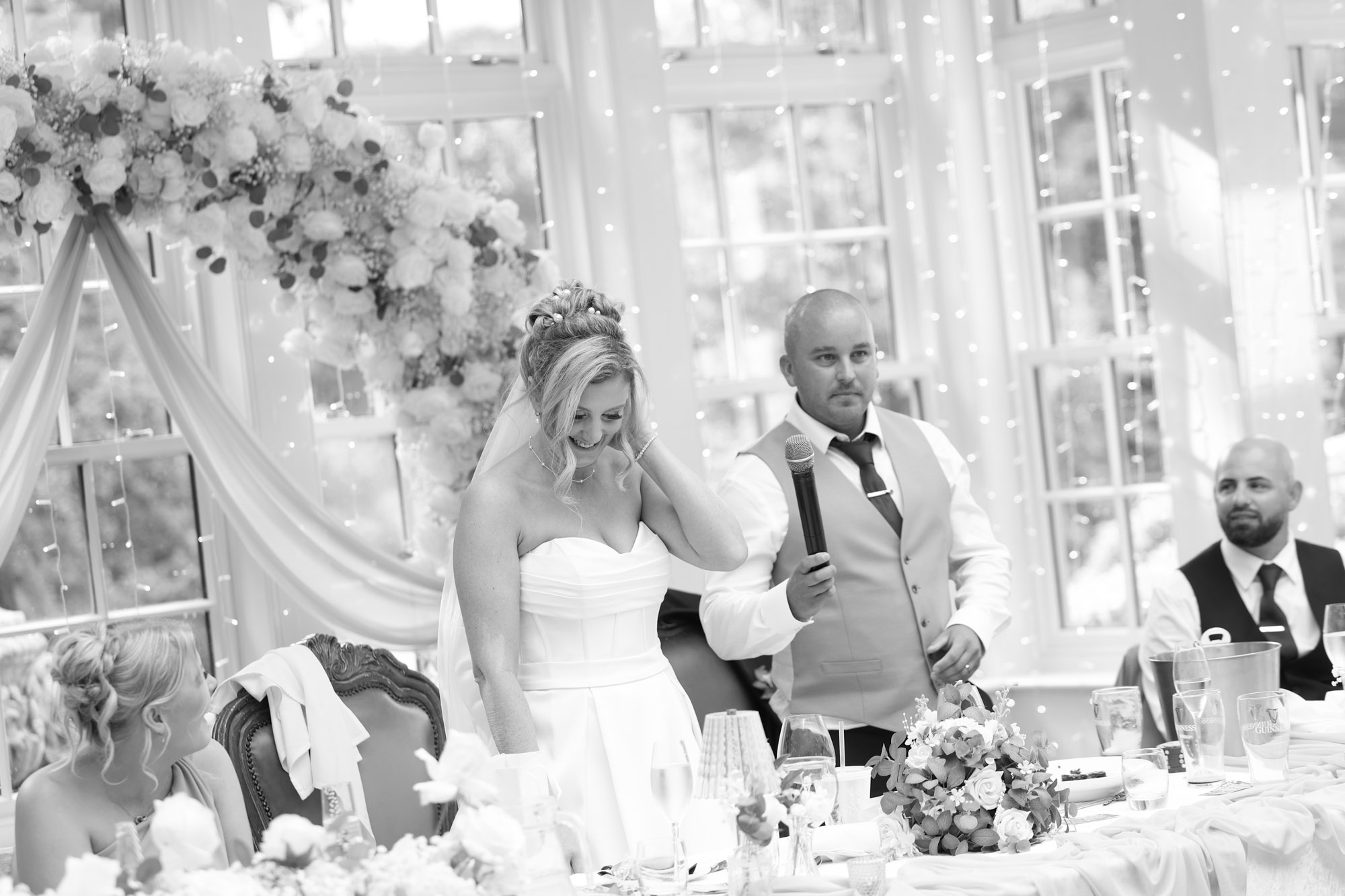 Black and white photo of the groom giving a speech with a microphone as the bride laughs at the top table at Mitton Hall.