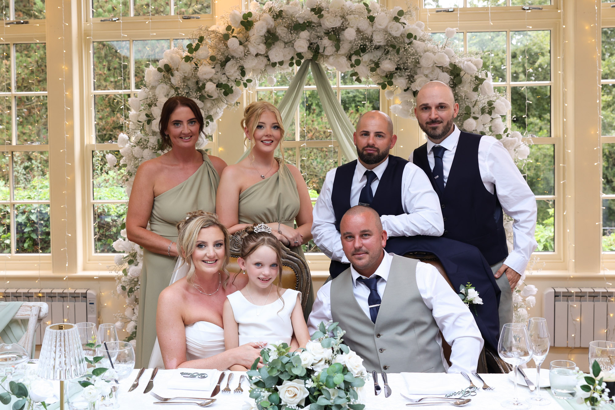 The wedding party posing together as part of a photo dash at the reception table in front of a floral arch at Mitton Hall.