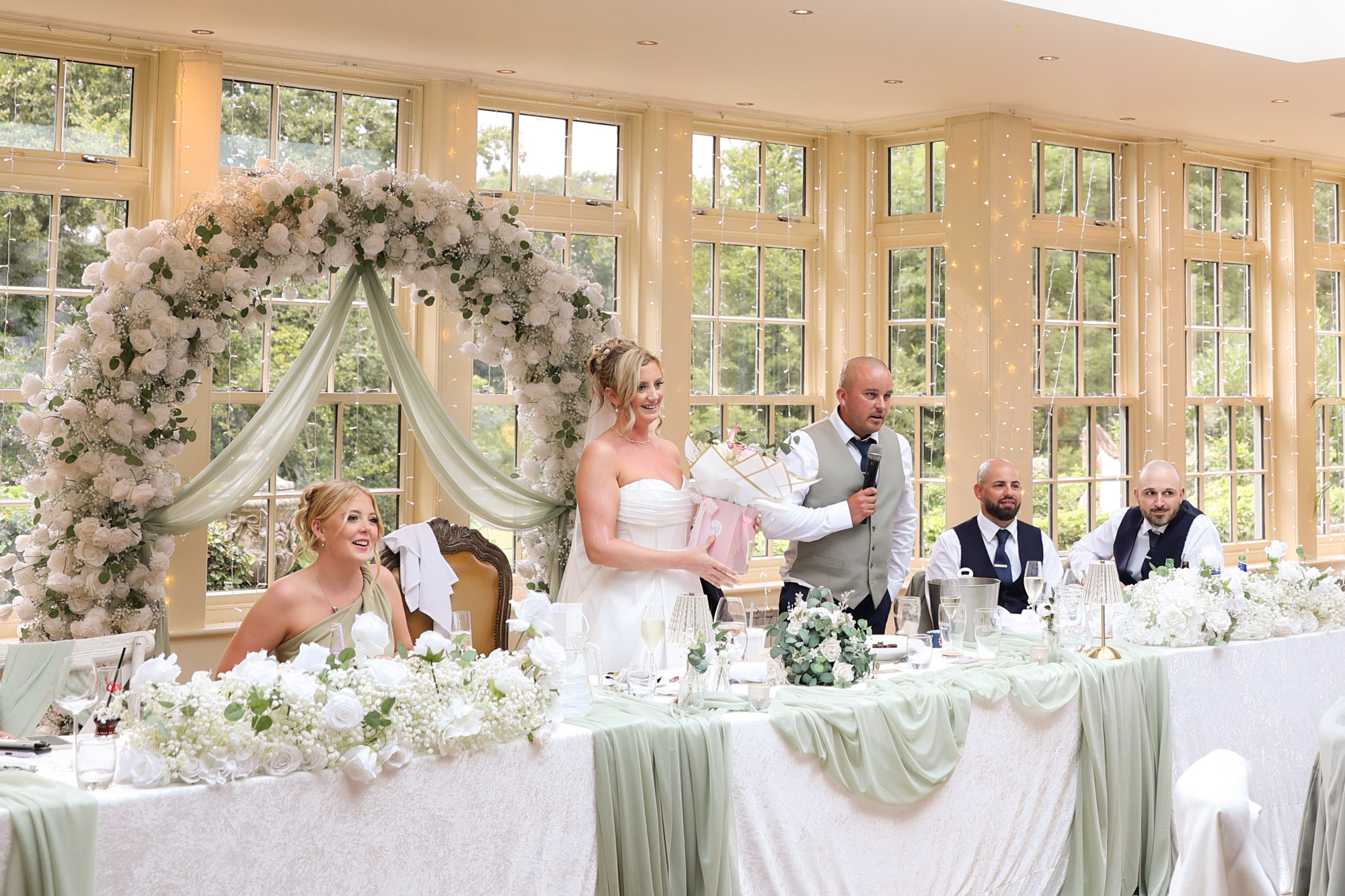 The groom speaking into a microphone while the bride holds a bouquet at the top table during a wedding reception at Mitton Hall.