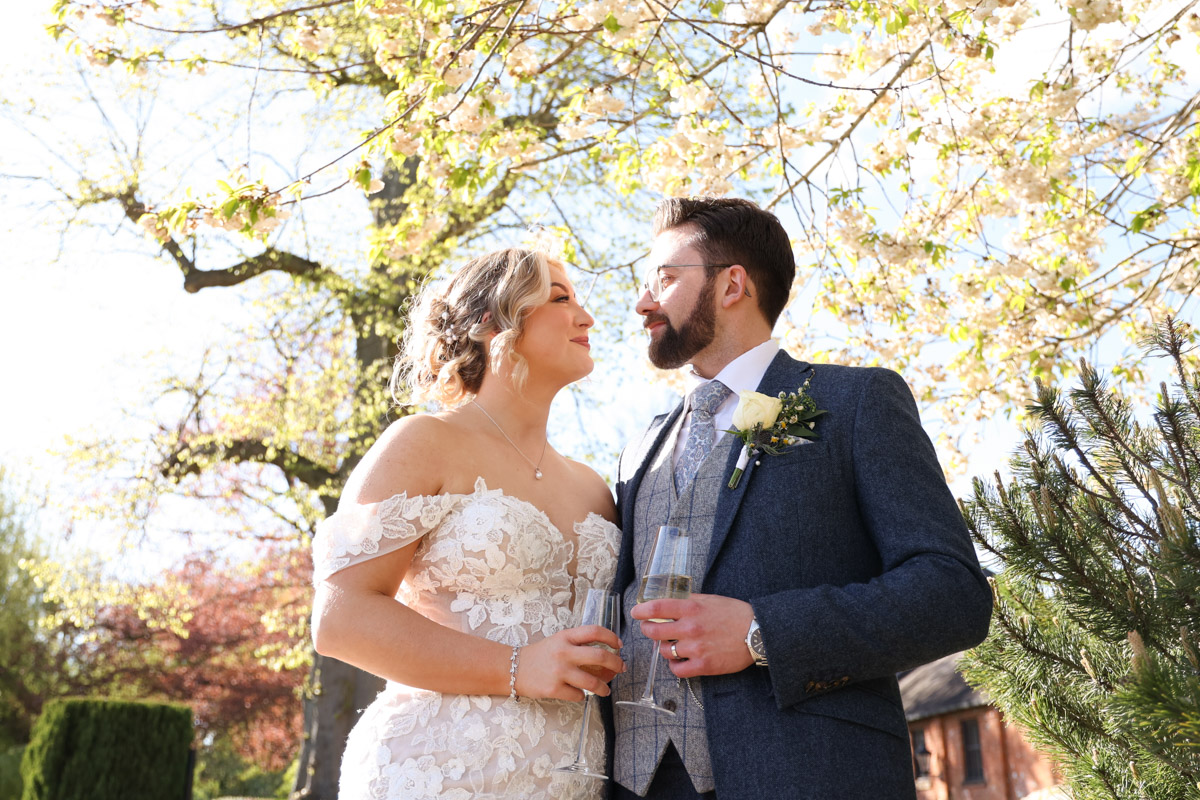 Bride and groom holding champagne flutes under blooming spring blossoms at Rookery Hall.