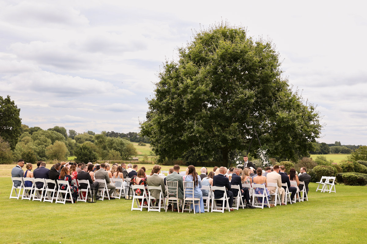 Guests seated on white folding chairs on a green lawn for an outdoor wedding ceremony at Rookery Hall, with a large tree in the background.