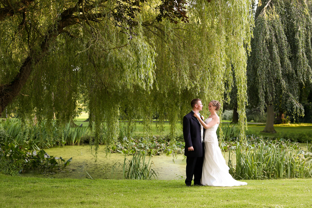 Newlywed couple standing under a weeping willow by a pond at Rookery Hall.