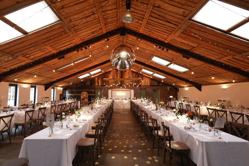 Interior of The Barn Owen House wedding barn set up for a reception with long banquet-style tables, white linens, wooden cross-back chairs, and a large disco ball hanging from the vaulted timber ceiling