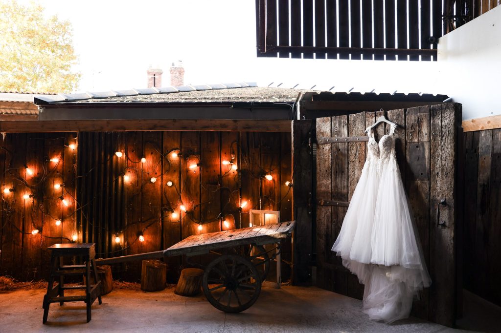A white lace wedding dress hangs on a large wooden door at The Barn Owl House; the rustic setting features a vintage wooden cart and a wall decorated with warm fairy lights