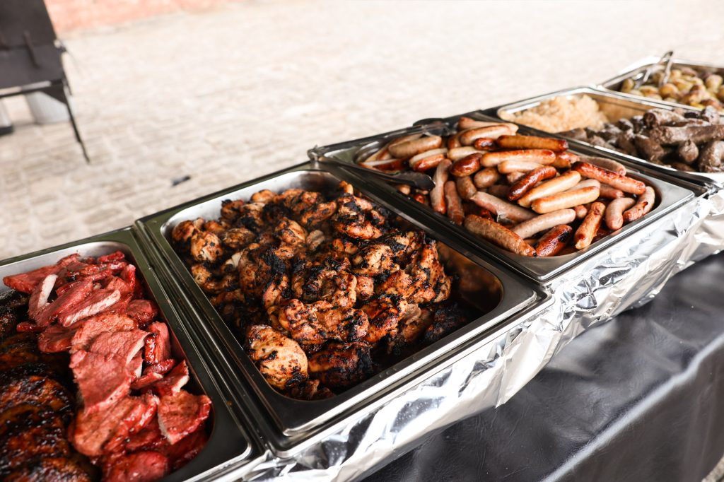 Catering buffet table with metal chafing dishes filled with roasted meats and sausages.