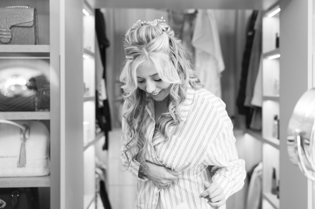 Black and white photo of a smiling blonde bride in a striped shirt inside a dressing area.