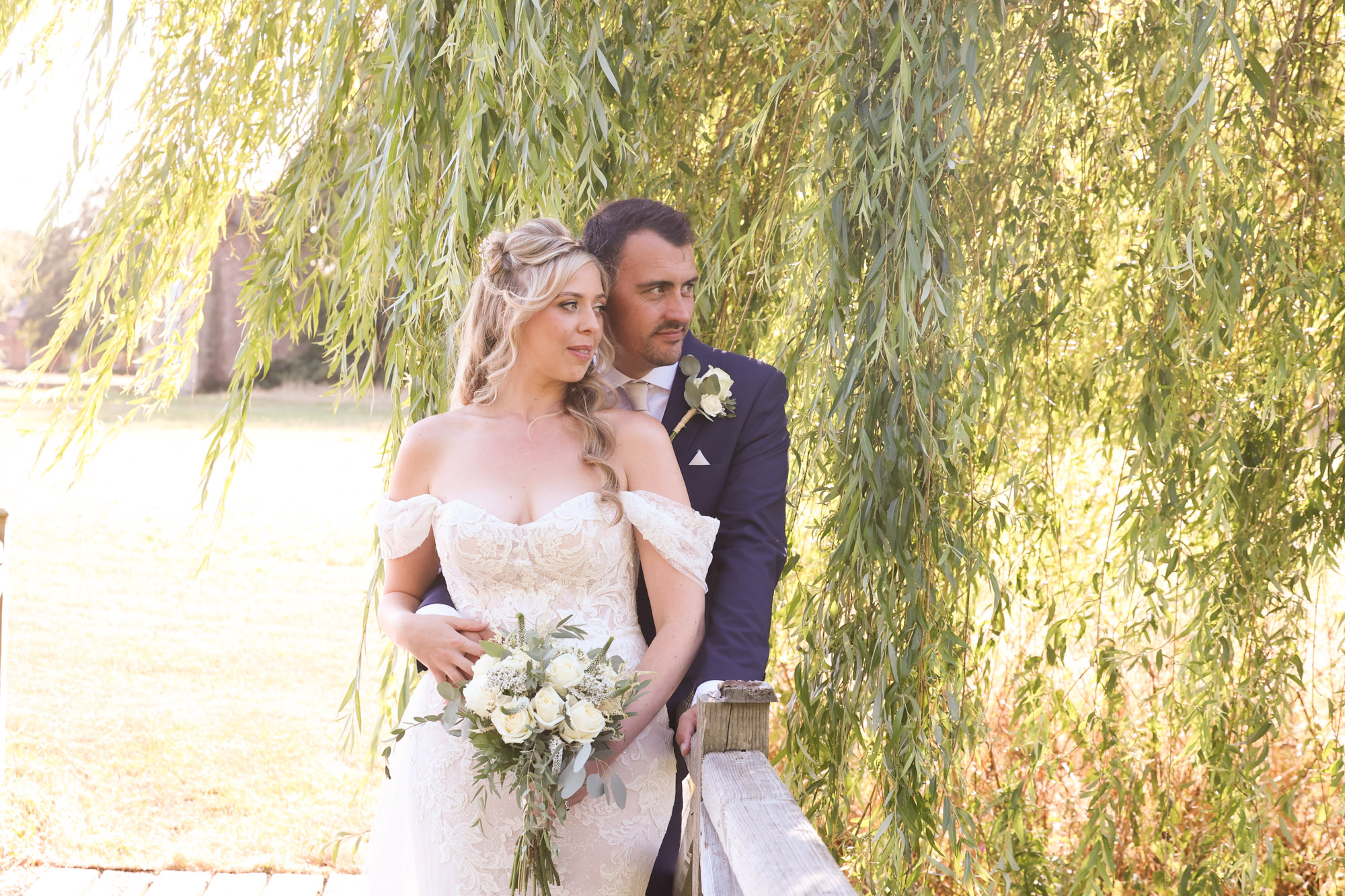 Bride and groom holding a bouquet, posing outdoors beneath weeping willow branches.