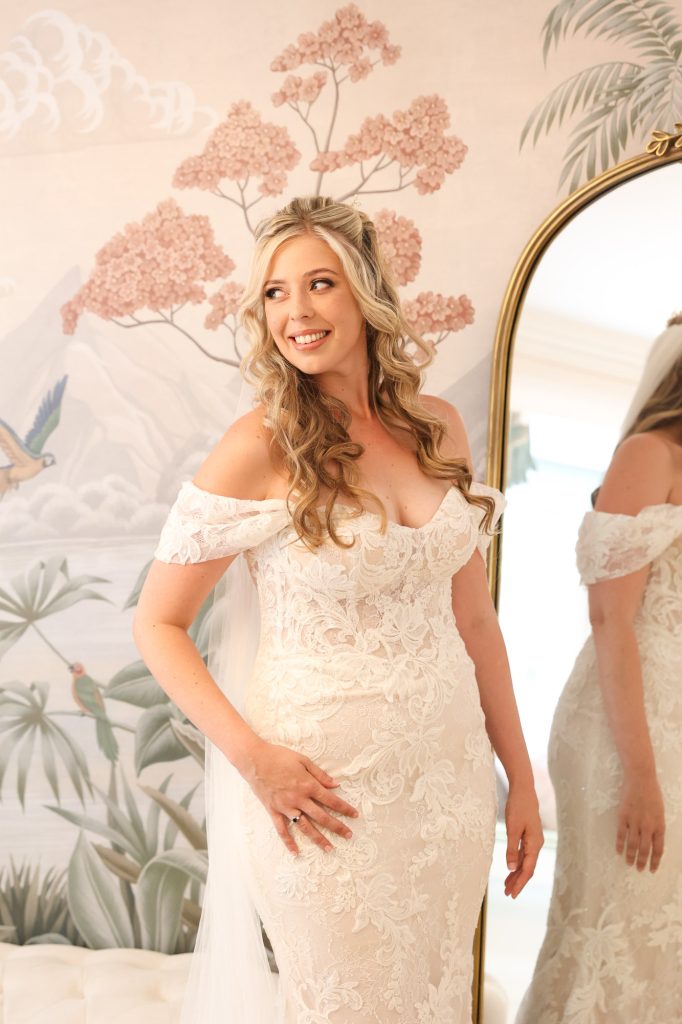 Bride smiling in a detailed lace gown against a floral wallpaper backdrop with a mirror.