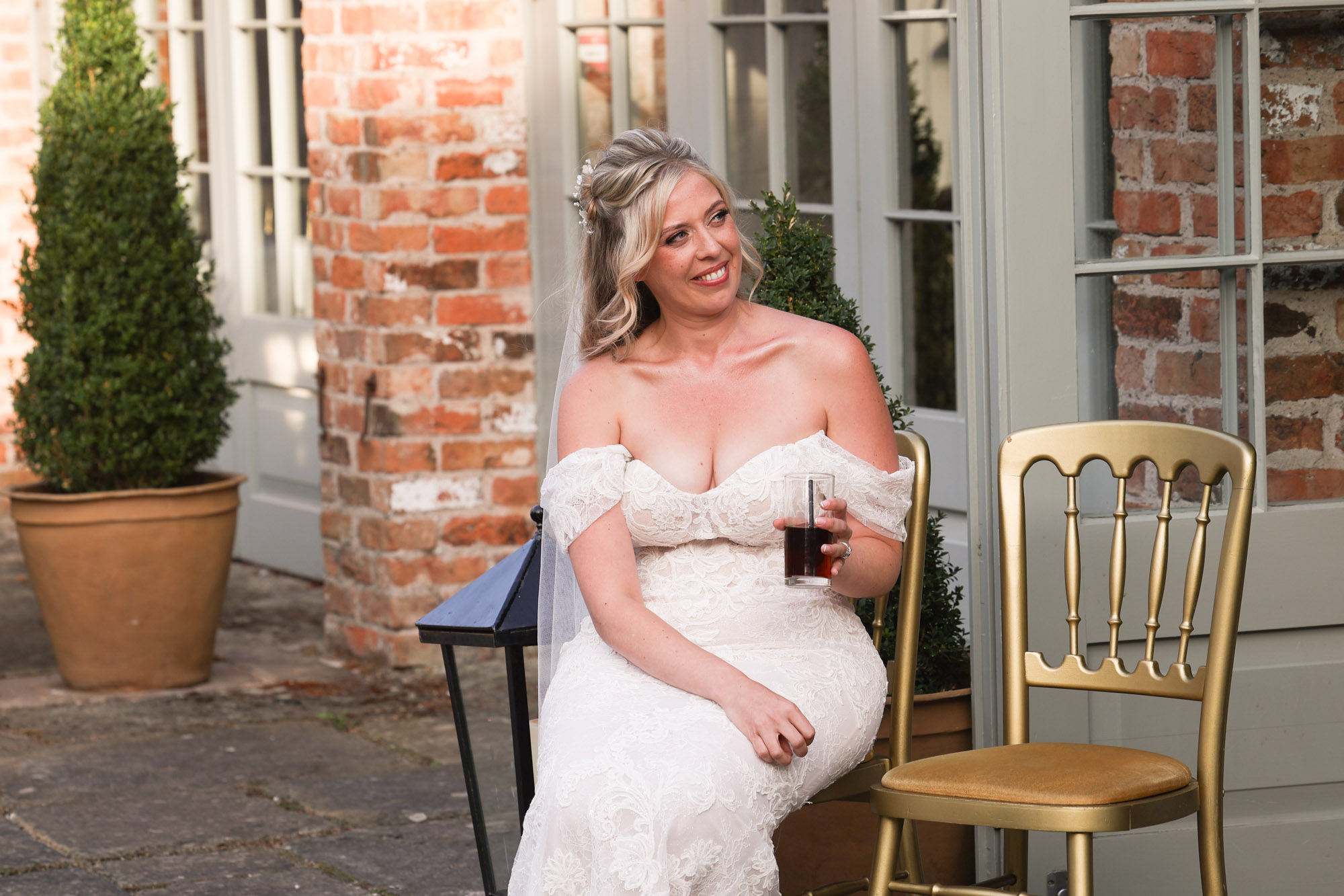 Bride in a white off-the-shoulder wedding dress holding a drink next to gold chairs.