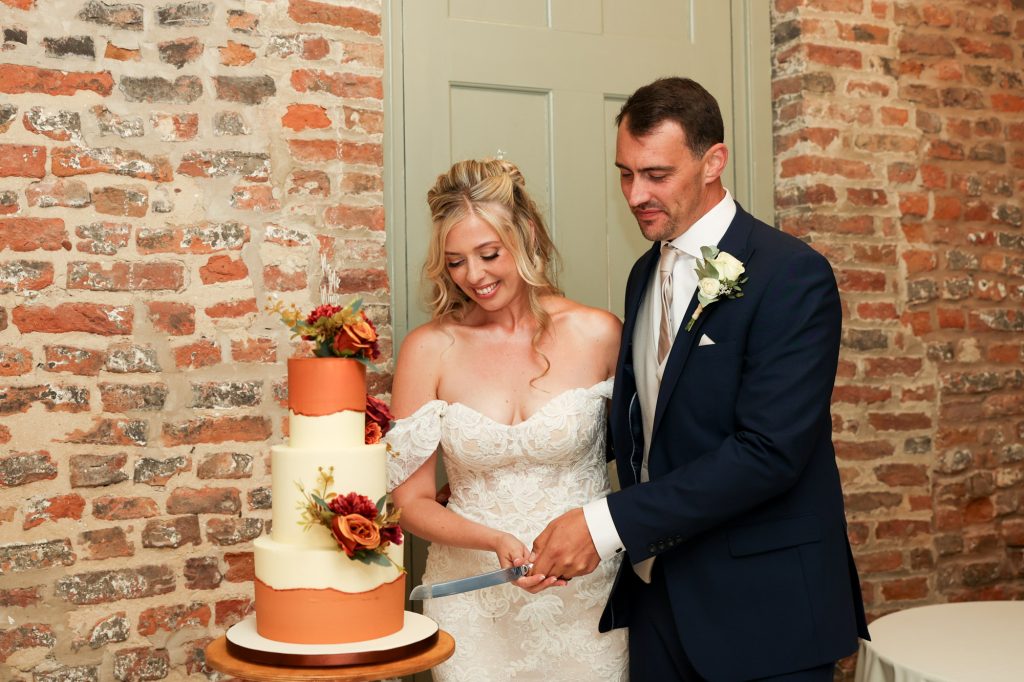 Bride and groom holding a knife together, cutting a three-tiered wedding cake decorated with autumnal flowers.