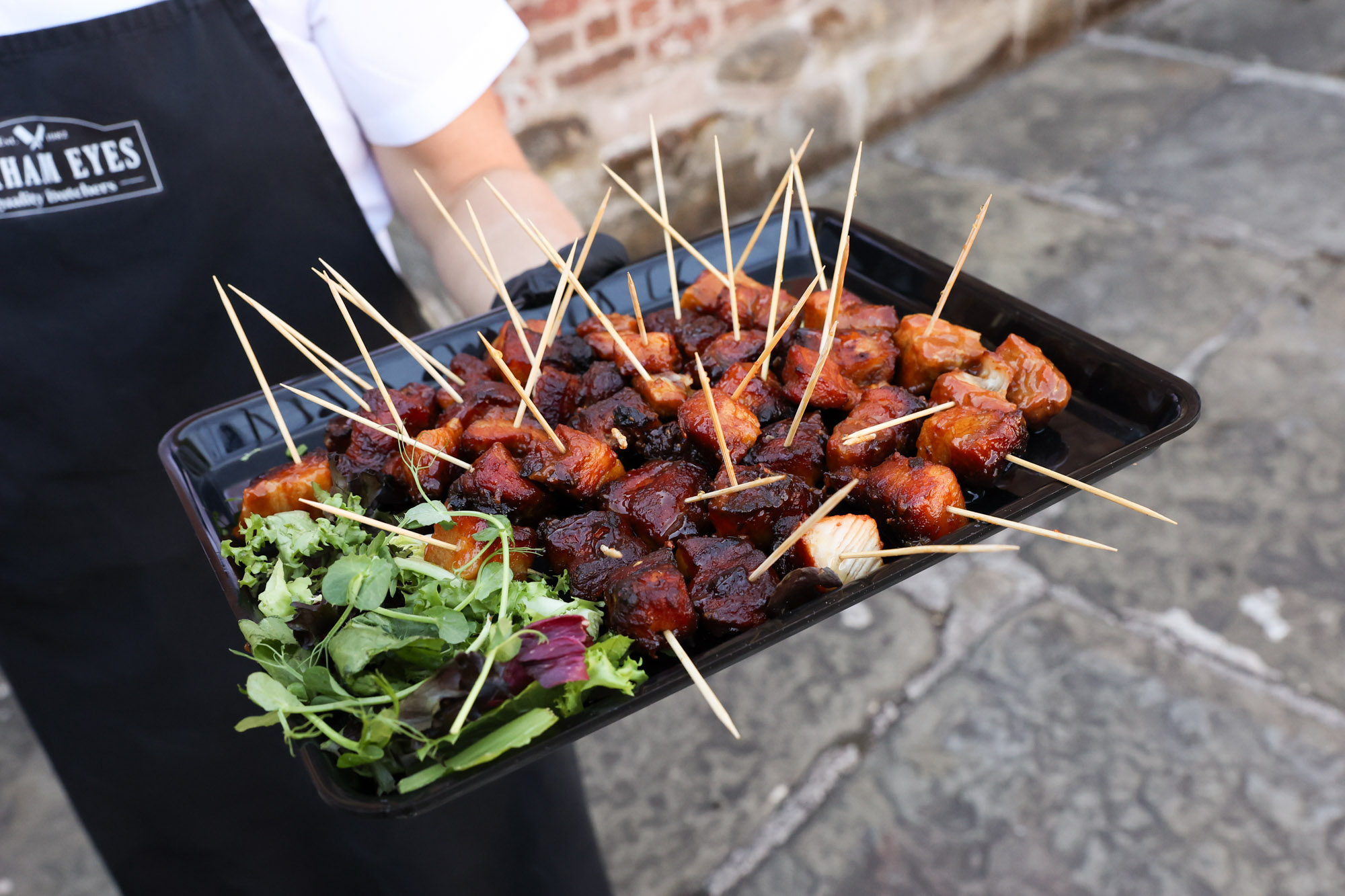 Close up of a server holding a black tray full of glazed meat skewers and a small salad garnish.