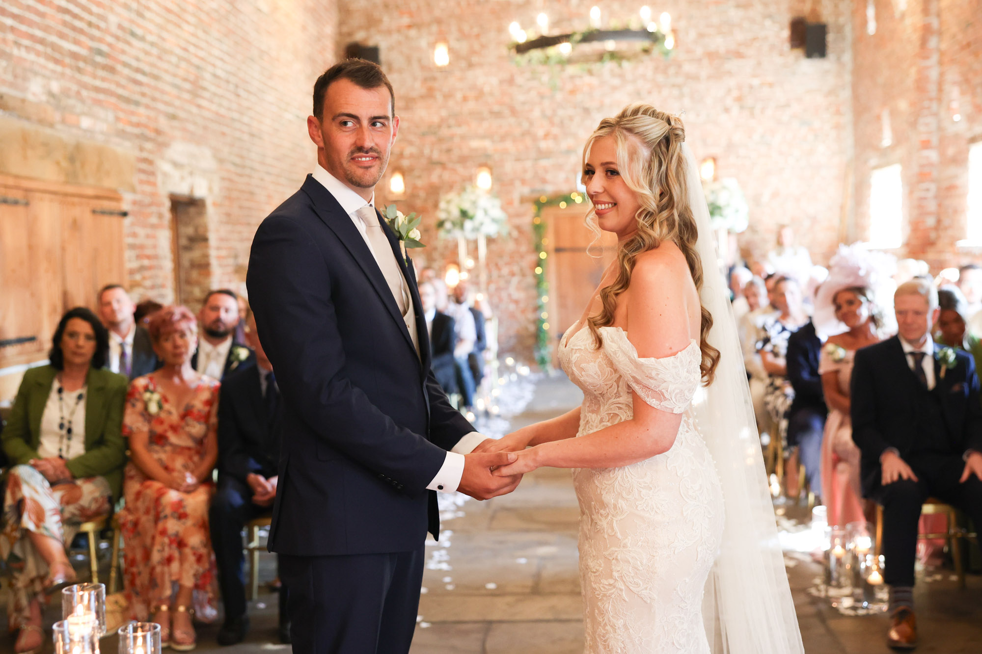 Bride and groom holding hands at the front of a rustic brick barn during their wedding ceremony, surrounded by guests.