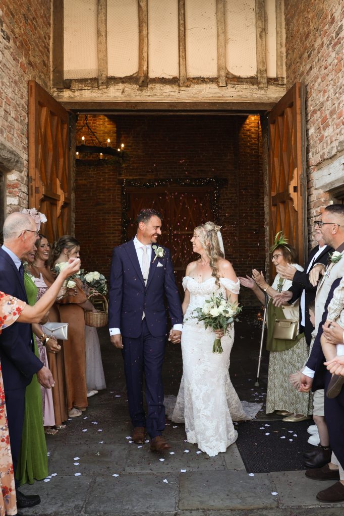 Smiling newlywed couple walking out of large wooden barn doors while guests throw confetti.